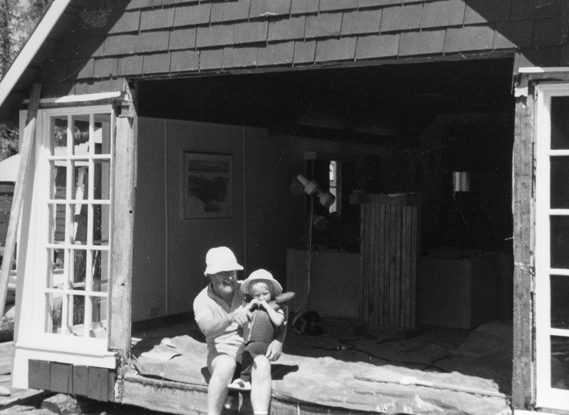 Man with beard sits holding a young child. Sitting on the side of a house cut open and inside exposed