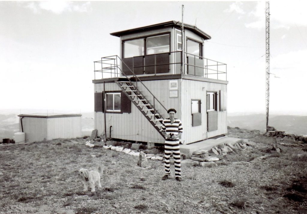 Man in a striped onesie standing with a dog in front of a fire lookout hut.